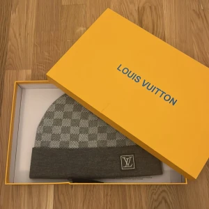 Grå rutig mössa från Louis Vuitton - Snygg grå mössa från Louis Vuitton med klassiskt rutmönster. Mössan har en uppvikt kant med LV-logotypen broderad på framsidan. Perfekt för att hålla stilen under kyliga dagar.