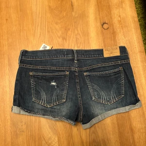 Mörkblå jeansshorts från Hollister - Säljer ett par mörkblå jeansshorts från Hollister med slitna detaljer och uppvikta kanter. Shortsen har en klassisk femficksdesign och låg midja. Perfekta för en avslappnad sommarlook!
