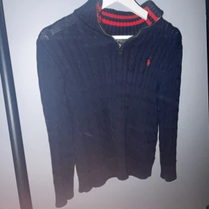 Mörkblå stickad tröja från Ralph Lauren - Säljer en snygg mörkblå stickad tröja från Ralph Lauren med dragkedja och röd logga på bröstet. Tröjan har en ribbad struktur och hög krage med röda ränder på insidan. Perfekt för kyliga dagar! Om de är nått ni undrar så fråga. Skulle säga att den är bra skick ungefär 8/10. Storlek 10/12 ungefär 146/152 