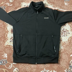 Svart jacka från Hugo Boss - Snygg svart jacka från Hugo Boss med dragkedja och vita detaljer längs ärmarna. Jackan har två praktiska fickor med dragkedjor och en stilren logga på bröstet. Perfekt för en sportig look.