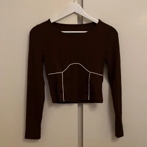 Brun långärmad topp med vita detaljer - OANVÄND - Snygg brun långärmad topp med vita kontrastlinjer vid brösten som ger en stilren look. Perfekt för en trendig outfit. Helt oanvänd!