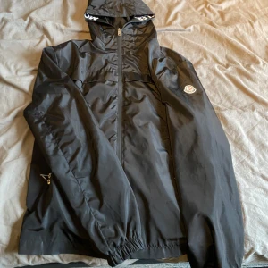Svart vindjacka från Moncler - Säljer en snygg svart vindjacka från Moncler. Jackan har en dragkedja framtill och en huva med logotypdetaljer. Perfekt för blåsiga dagar och har en stilren look. Passar både till vardags och lite finare tillfällen.