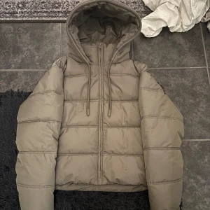 Grön pufferjacka med huva - Säljer en snygg grön pufferjacka med huva. Jackan har en dragkedja framtill och snörning vid huvan för extra skydd mot kyla. Perfekt för höst och vinter, håller dig varm och skyddar mot vind. Jackan är i nyskick och helt oanvänd. Säljer pga att jag egentligen ville lämna tillbaks men hade dragit av lappen. Köptes för 559kr men säljer för 500kr. Storlek XS 