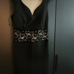 Svart klänning från Bodyflirt - Elegant svart klänning från Bodyflirt med spetsdetaljer i midjan. Den har en v-ringad design och är ärmlös, vilket gör den perfekt för festliga tillfällen. Klänningen är i nyskick och har en figurnära passform.