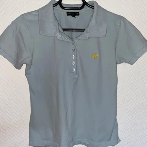 Grå polotopp från H&M - Säljer en stilren grå polotopp från Ellos i bra skick. Den har korta ärmar och en klassisk krage med knappar framtill. Perfekt för både vardag och lite mer uppklädda tillfällen. Den lilla gula broderade kronan på bröstet ger en extra touch. Frågor i dm💌