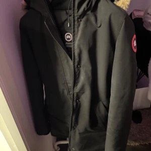 Svart vinterjacka från Canada Goose - Säljer en svart vinterjacka från Canada Goose i storlek M. Jackan har en pälsfodrad huva och är perfekt för kalla vinterdagar. Den har en dragkedja och knappar framtill samt det klassiska Canada Goose-märket på ärmen. Supervarm och stilren! Den behöver tvättas!!