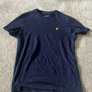 Mörkblå t-shirt från Lyle & Scott - Säljer en snygg mörkblå t-shirt från Lyle & Scott i mycket bra skick. Den har en gul broderad logga på bröstet och är tillverkad i mjuk bomull. Perfekt för både vardag och fest! Passar bra till jeans eller shorts.