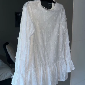 Vit klänning NA-KD med blomdetalj - Säljer denna vita korta klänning från NA-KD i storlek 36. Den har knyte i ryggen. Jättefin att ha på studenten eller sommaren!!