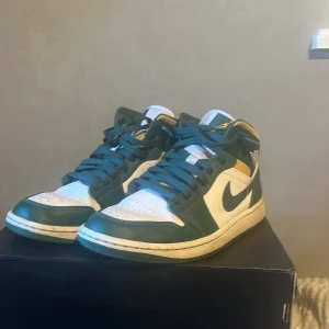 Nike Air Jordan 1 Mid - Säljer ett par Nike Air Jordan 1 Mid i grönt och vitt. Skorna har en klassisk design med snörning och är i bra skick. Perfekta för både vardag och festliga tillfällen. De är bekväma och stilrena, en riktig ikon i sneaker-världen!