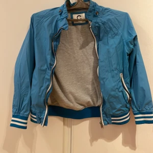 Blå vindjacka för barn - Säljer en snygg blå vindjacka för barn från märket C. Jackan har en dragkedja framtill och ribbade muddar vid ärmslut och nederkant. Perfekt för vår och höst! Passar barn i åldern 8 år. 🌀