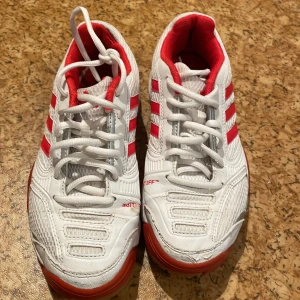 Vita och röda sneakers från Adidas - Säljer ett par snygga vita sneakers från Adidas med röda detaljer och snörning. Skorna har en sportig look med tre röda ränder på sidan och en bekväm passform. Perfekta för träning eller vardagsbruk!