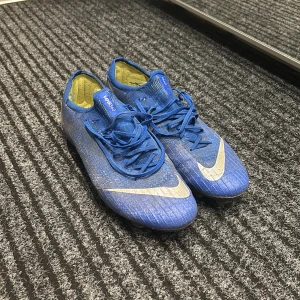 Blå Nike Mercurial fotbollsskor - Säljer ett par blå Nike Mercurial fotbollsskor i bra skick. De har en snygg design med vita detaljer och snörning. Perfekta för fotbollsträning eller match. Skorna har använts men är fortfarande i gott skick med bra grepp på dobbarna.