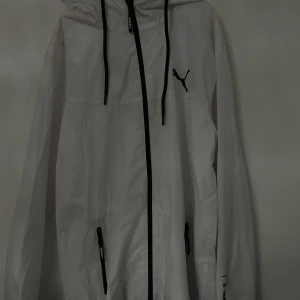 Vit vindjacka från Puma - Säljer en snygg vit vindjacka från Puma. Den har en dragkedja framtill och justerbar huva med svarta snören. Jackan är perfekt för vår och höst, och har två praktiska fickor med dragkedja. Den är i mycket bra skick och passar perfekt för en sportig look. S/M