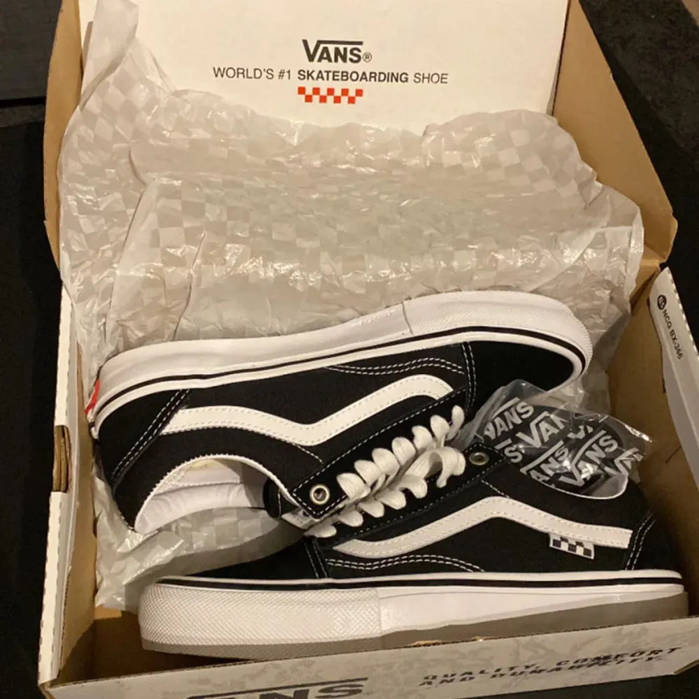 Säljer ett par klassiska svarta Vans sneakers i nytt skick. De har den ikoniska vita sidostripen och checkerboard-mönster på insidan. Perfekta för skateboarding eller vardagsbruk. Storlek är inte specificerad, men de ser ut att vara i standard passform. Kommer i originalkartong.. Skor.