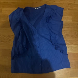 Blå blus från WE - Säljer en snygg blå blus från WE i storlek S. Den har en v-ringad design med knappar framtill och volangdetaljer längs ärmarna. Perfekt för både vardag och fest!
