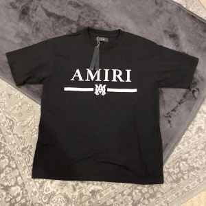 Svart t-shirt från Amiri - Säljer en snygg svart t-shirt från Amiri i nytt skick. Den har ett stilrent tryck med 'AMIRI' i vitt på framsidan. Perfekt för en casual look eller att klä upp med en snygg jacka. T-shirten är kortärmad och gjord i mjukt bomullsmaterial. Passar perfekt för både vår och sommar!