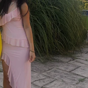 Ljusrosa klänning med volangdetaljer - Säljer en superfin ljusrosa klänning med volanger och en elegant slits. Perfekt för sommarens fester eller en romantisk kväll. Klänningen är axelbandslös och har en tight passform som framhäver figuren. Den är både söt och stilren, och passar perfekt för en festlig look!