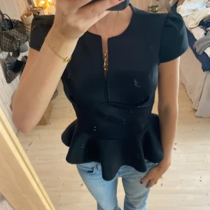 Svart peplumtopp - Snygg svart peplumtopp med korta ärmar och en liten v-ringning. Perfekt för en kväll ute eller en fest. Toppen har en figursydd passform som framhäver midjan och ger en elegant siluett. Materialet känns bekvämt och stilrent.