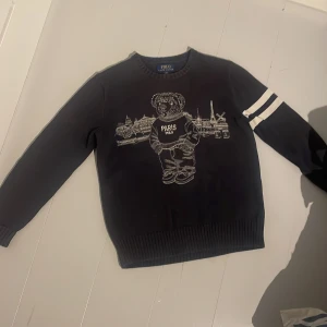 Säljer en snygg mörkblå stickad  Ralph lauren tröja, polo paris. Väldigt fint skick på tröjan. Tröjan är perfekt nu till vinter. Den passar dig fint om man är ungefär mellan 155-168. - Säljer en svart tröja från Polo med ett coolt Paris-motiv och en nalle på framsidan. Tröjan har långa ärmar med två vita ränder på ena ärmen. Perfekt för höst och vinter, och den är i bra skick. Passar både till vardags och lite mer avslappnade tillfällen.
