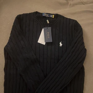 Svart stickad tröja från Ralph Lauren - Säljer en snygg svart stickad tröja från Ralph Lauren. Den har ett klassiskt kabelstickat mönster och den ikoniska loggan broderad på bröstet. Perfekt för både höst och vinter, och den är i helt nytt skick med taggar kvar. Passar till alla tillfällen!