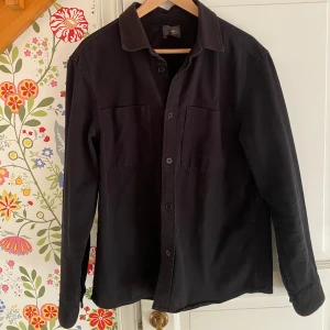Svart overshirt från H&M - Säljer en stilren svart overshirt från H&M i storlek M. Perfekt för lager-på-lager under höst och vår. Skjortan har långa ärmar och knappar framtill, vilket ger en klassisk look. Materialet känns slitstarkt och bekvämt. Passar både till vardags och lite mer uppklädda tillfällen.