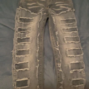 Grå jeans med slitningar från Amiri - Säljer ett par grå jeans från Amiri med massor av coola slitningar längs benen. De har en edgy och trendig look, perfekt för en avslappnad stil. Jeansen är i bra skick och har en knappstängning framtill.