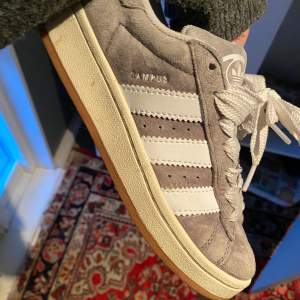 Säljer ett par grå Adidas Campus sneakers i mocka med vita detaljer och snörning. De har en klassisk design med tre vita ränder på sidan och en gummisula i beige. Skorna är i bra skick och kommer med extra skosnören i både grått och vitt. Storlek 36