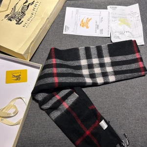 Burberry halsduk - Säljer en elegant rutig halsduk från Burberry i svart, vit och röd. Den är i ull och har en klassisk design som passar perfekt för höst och vinter. Halsduken är i nytt skick och kommer med originalförpackning och kvitto. Perfekt för att hålla sig varm med stil!