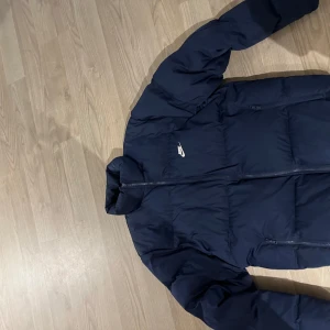 Mörkblå puff jacka från Nike - Säljer en snygg mörkblå dunjacka från Nike i mycket bra skick. Jackan har en dragkedja framtill och är perfekt för kalla dagar. Den är långärmad och har en normal passform. Perfekt för höst och vinter!