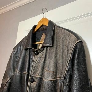 Vintage Svart skinnjacka Paramount Pictures - Säljer en svart skinnjacka från Paramount i äkta läder. Jackan har en klassisk stil med knappar framtill och två innerfickor, två yttre. Perfekt för höst och vår. Den är i riktigt solblekt med en vacker patina och har en snygg vintage-look. Själva jackan är i väldigt bra skick, bara solblekningen. 