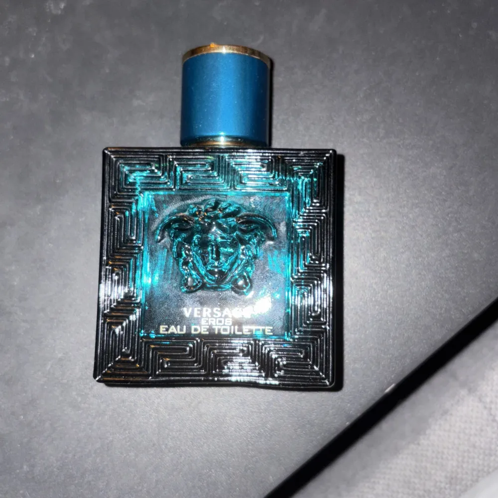 Säljer en flaska Versace Eros Eau de Toilette. Flaskan är fyrkantig och mörkblå med ett snyggt mönster och Versaces ikoniska Medusa-logga på framsidan. Korken är också blå med en gyllene topp. Perfekt för den som vill ha en fräsch och maskulin doft. Passar både till vardags och fest!. Perfume.