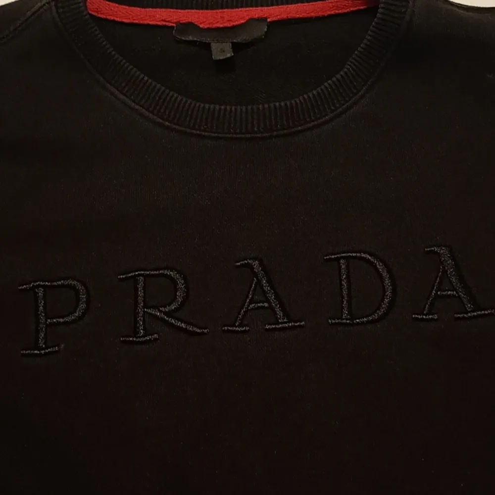 Säljer en stilren svart tröja från Prada med broderad logga på bröstet. Tröjan har långa ärmar och en röd detalj vid kragen. Perfekt för både vardag och fest!. Tröjor & Koftor.