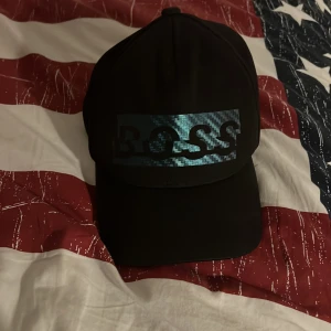 Svart keps från Hugo Boss - Säljer en stilren svart keps från Hugo Boss med en cool, glansig logga framtill. Kepsen har justerbar rem 