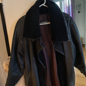 Svart skinnjacka med pälsfoder från Friitala - Säljer en snygg svart skinnjacka från Friitala med mjukt pälsfoder. Jackan har en klassisk design med dragkedja och en stor krage i ull. Vintage 