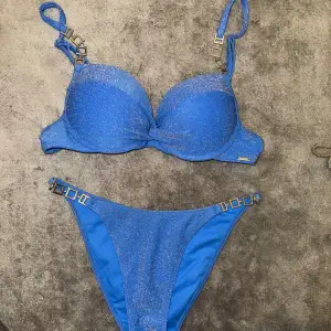 Snygg blå bikini med glittrigt material och dekorativa spännen på axelbanden och sidorna av trosorna. Perfekt för sommarens stranddagar! Bikinin har en elegant design med en twist-detalj på toppen. Den är både stilren och iögonfallande. Toppen passar en 34 a i byst underdelen är xs bikinitoppen har puchup 