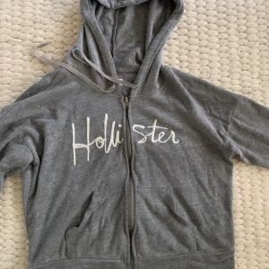 Grå hoodie från Hollister - Säljer en grå hoodie från Hollister i storlek XS. Den har en dragkedja framtill och en mysig huva. Perfekt för en avslappnad stil eller kyliga dagar. Hollister-loggan är insytt i vitt på framsidan. Skön och stilren!