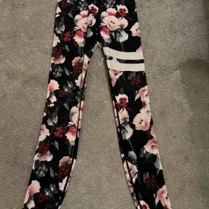 Blommiga leggings från Stronger - Säljer ett par snygga blommiga leggings från Stronger i storlek XXS. De är svarta med ett vackert blommönster i rosa och rött. Perfekta för träning eller vardagsbruk. Materialet är 85% återvunnen polyester och 15% elastan, vilket gör dem både bekväma och stretchiga.