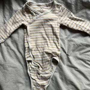 Randig omlottbody från Lindex - Säljer en superfin randig omlottbody från Lindex i storlek 68. Den är vit med gula ränder och har långa ärmar. Perfekt för både vår och höst! Den är i bra skick och har praktiska knappar för enkel påklädning. Perfekt för din lilla stjärna! 🌟