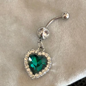 Navelpiercing med grönt hjärta och strass  - Snygg navelpiercing med ett stort grönt hjärta omgivet av glittrande vita stenar. Helt ny! Den har en böjd stav med en klar sten på toppen. Kirurgisk stål oanvänd 