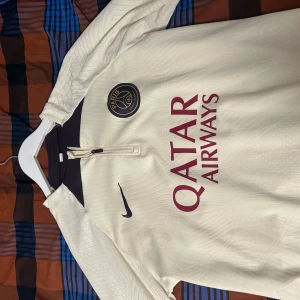 PLAYER EDITION PSG TRÖJA 23/24 - Säljer en snygg beige fotbollströja från Nike med PSG-logga och Qatar Airways sponsortryck. Tröjan har korta ärmar och en stilren design med mörka detaljer vid kragen. Perfekt för fotbollsfans som vill ha något unikt i garderoben! 🏆byxa går att Få med 