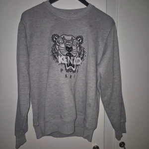 Grå sweatshirt från Kenzo - Säljer en snygg grå sweatshirt från Kenzo med deras ikoniska tigerbroderi på framsidan. Tröjan är i bra skick och har långa ärmar. Perfekt för en casual look eller att mysa i hemma. Passar både höst och vår!