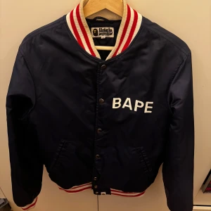 Mörkblå bomberjacka från BAPE - Säljer en snygg mörkblå bomberjacka från BAPE med vita och röda detaljer. Jackan har ett coolt tryck med texten 'A Bathing Ape' på ryggen och 'BAPE' på framsidan. Den är quiltad inuti för extra komfort och värme. Perfekt för höst och vår!