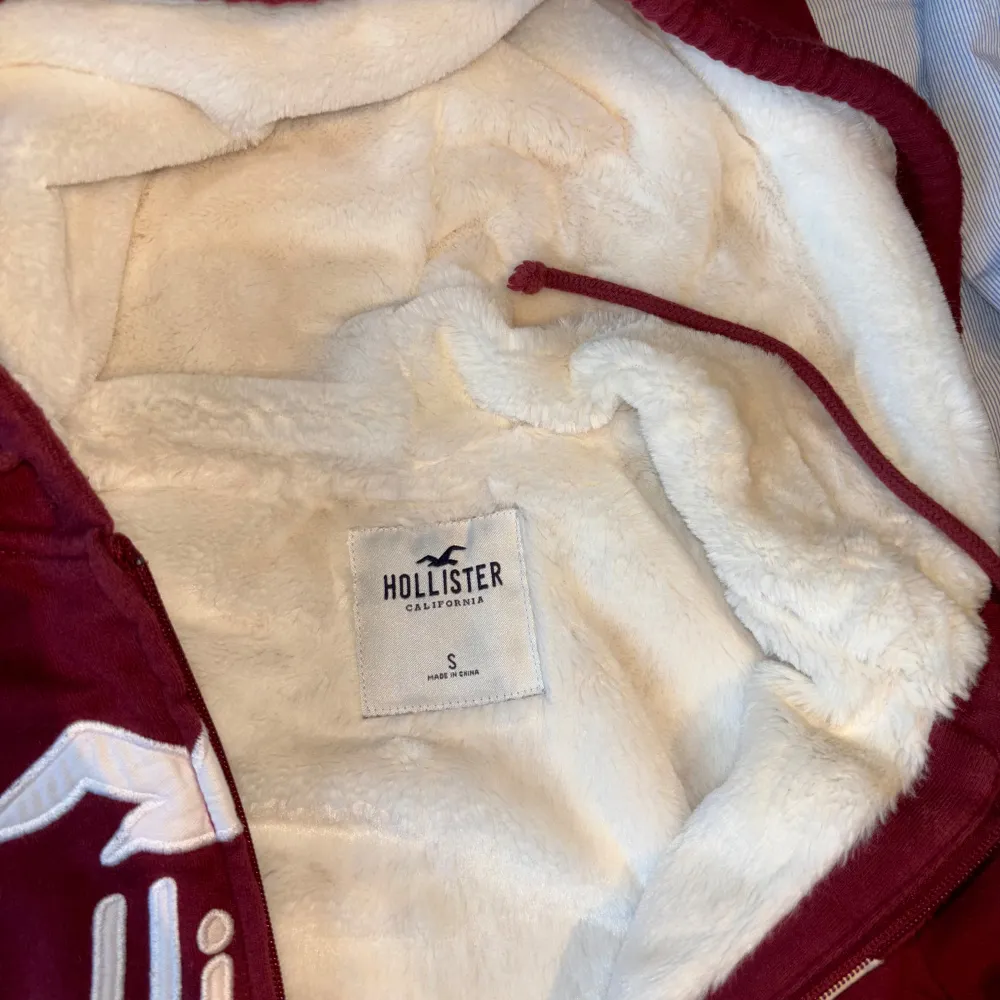 Säljer en supermysig röd hoodie från Hollister med vit text och logga på framsidan. Den har en dragkedja och är fodrad med mjuk, vit fluff på insidan. Perfekt för kyliga dagar när du vill vara både varm och stilren. 🧥. Hoodies.