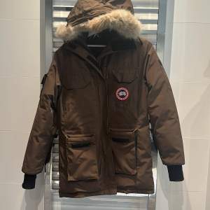 ÄKTA FRÅN LÄNNA SPORT Säljer en brun parkajacka från Canada Goose, perfekt för kalla vinterdagar. Jackan har en avtagbar huva med pälsdetaljer och flera praktiska fickor. Den är långärmad och har en dragkedja framtill. Supervarm och stilren!