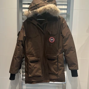 Brun parkajacka från Canada Goose - ÄKTA FRÅN LÄNNA SPORT Säljer en brun parkajacka från Canada Goose, perfekt för kalla vinterdagar. Jackan har en avtagbar huva med pälsdetaljer och flera praktiska fickor. Den är långärmad och har en dragkedja framtill. Supervarm och stilren!