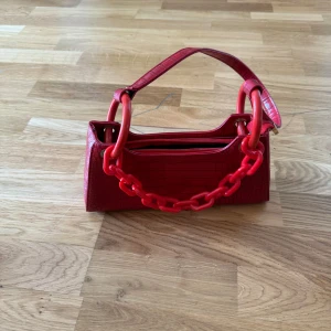 Röd axelväska med kedjedetalj - Säljer en supercool röd axelväska med en chunky kedjedetalj. Väskan är i syntetmaterial med en krokodilpräglad yta och har en dragkedja upptill. Perfekt för att ge din outfit en färgklick! 💥