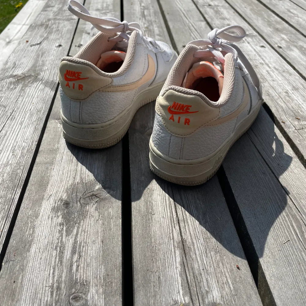 Säljer ett par klassiska Nike Air Force sneakers i vitt med orange detaljer. Skorna har en snygg och stilren design med snörning och en bekväm passform. Perfekta för både vardag och fest! Stl 37,5 Dam. Skor.