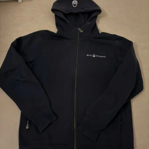 Sailracing hoodie - Säljer denna marinblå sailracing hoodie i ett bra skick. Saknas snören vid luvan och det är den ända defekten. Den är i en herrstorlek S men funkar bra till dam med. 