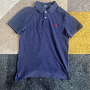 Mörkblå pikétröja från Ralph Lauren - Säljer en snygg mörkblå pikétröja från Ralph Lauren. Den har en klassisk krage med knappar och det ikoniska röda broderade logotypen på bröstet. Perfekt för en avslappnad men stilren look!