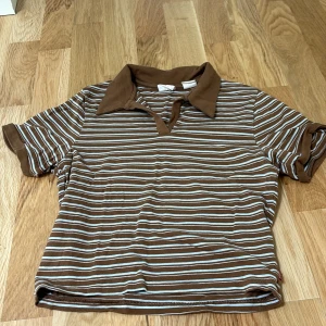 Brun och vit randig pikétröja från Levi's - Säljer en snygg brun och vit randig pikétröja från Levi's i storlek XS. Tröjan har korta ärmar och en klassisk krage. Perfekt för en avslappnad stil, och den är i jätte bra skick, ej använt. original pris 400kr